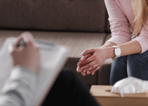 A light skin toned woman sits in a therapy session. You see her hands and the therapists hands. The therapist is writing on a notepad.