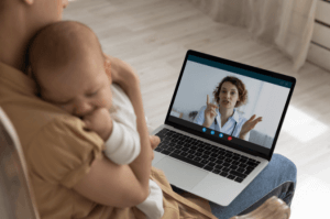 A new mother holds her sleeping baby against her chest. The mother has her laptop resting on her laptop and is in a video chat meeting.