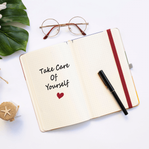 A white notebook, a pair of glasses, and a green leaf on a white surface. The notebook says take care of yourself and has a red heart drawn on it.