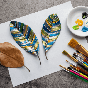 Two decorated leaves with colorful patterns next to a palette of paint and various brushes on a textured grey background. Art projects during a thunderstorms can help relax a child experiencing child anxiety about weather. Look for a child therapist in Branchburg or Scotch Plains today!