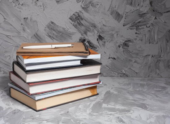 A stack of colorful books with a notebook and pen on top, set against a gray textured background. Learn more about overcoming back to school anxiety by searching for child counseling in Branchburg, NJ. Contact a child therapist in Branchburg, NJ to learn more about child counseling Scotch Plains, NJ and other services.