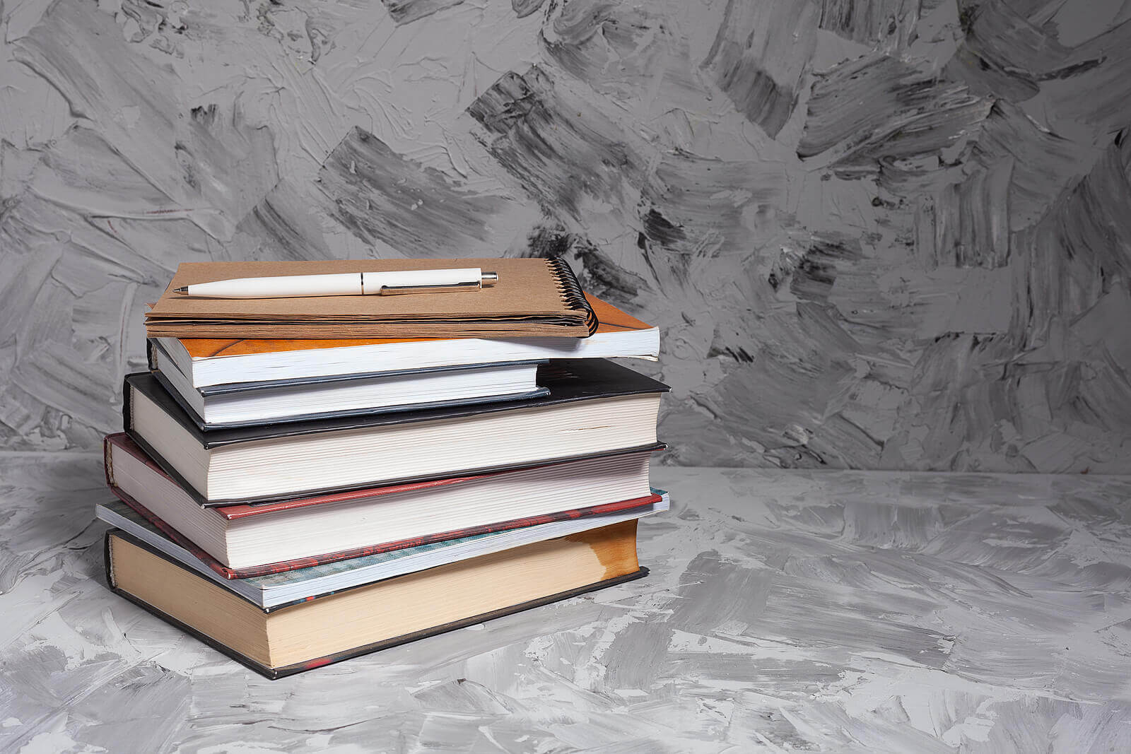 A stack of colorful books with a notebook and pen on top, set against a gray textured background. Learn more about overcoming back to school anxiety by searching for child counseling in Branchburg, NJ. Contact a child therapist in Branchburg, NJ to learn more about child counseling Scotch Plains, NJ and other services.