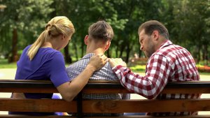 A mother and father sit on a park bench, on either side of their teen child. They talk with serious expressions, representing a hard conversation that therapy for teens in Branchburg, NJ can help address. Learn more about online therapy for teens by searching for counseling for teens in Scotch Plains, NJ today.