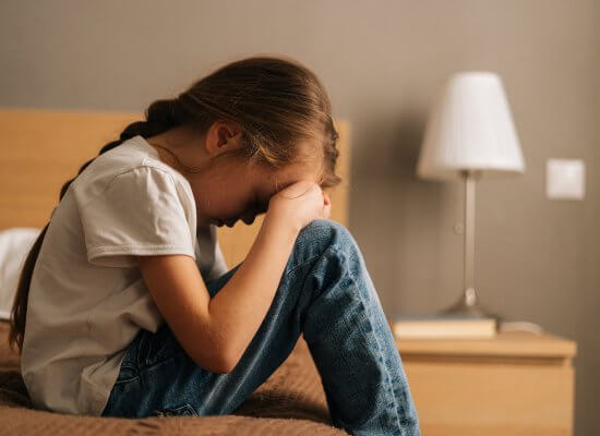 A child sits on a bed with her head resting on her knees. This could represent the isolation of anxiety that a child therapist in Branchburg, NJ can help address. Learn more about anxiety treatment in Branchburg, NJ or child therapy for anxiety. Search for an anxiety therapist in Branchburg, NJ today.