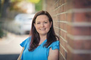 A woman with long dark hair in a blue shirt is smiling while leaning against a brick wall outside. She is a selective mutism therapist with Brave Minds Psychological Services. Learn more about selective mutism in Branchburg, NJ and how a selective mutism therapist in Somerville, NJ can help today.