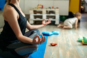 Person meditating on a yoga mat in a room while a child crawls on the floor nearby.