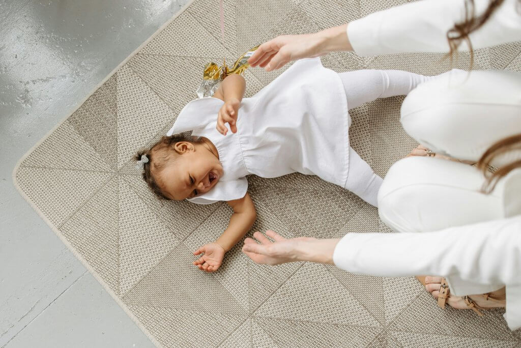 Photograph of a baby on a carpet with the mothers hands reaching out to them