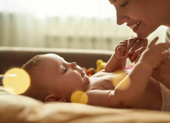 A woman smiles at a baby lying on a bed, holding its hands. The background is softly lit with warm tones.