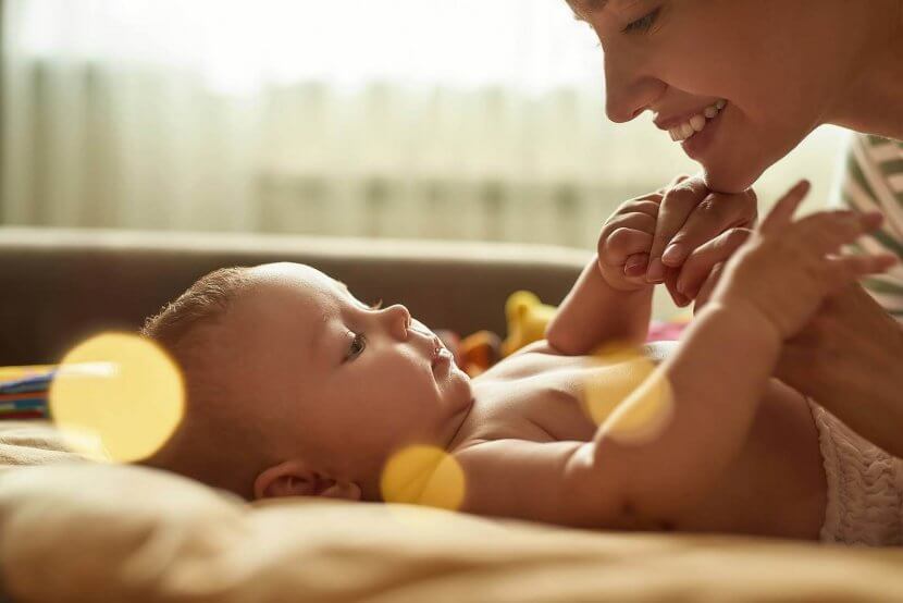 A woman smiles at a baby lying on a bed, holding its hands. The background is softly lit with warm tones.