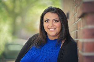 A person with long dark hair smiles, wearing a blue top and black cardigan, leaning against a brick wall, with a blurred green background. Learn how an LGBTQIA+ therapist in Branchburg, NJ can help you in finding a supportive, safe place. Search for LGBTIQIA+ affirmative therapy in Somerville, NJ today.