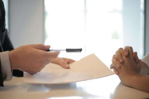 Two people at a table. One, a therapist, holds a pen and a document, while the other has hands clasped thoughtfully. A bright window in the background casts gentle light across their conversation. This could be a life-changing conversation for those looking for the right diagnosis. Contact us to today to learn more.