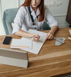 A neuroaffirming therapist near Somerville, NJ in a white coat writes in a notebook at a wooden table adorned with a tablet, smartphone, pencil, and blister packs of pills. Find a therapist who understands your struggle. Call us today.