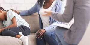 A child faces away from their parent while sitting on a sofa across from a woman with a clipboard. This could represent the support a selective mutism therapist can offer in addressing selective mutism in Branchburg, NJ. Search for childhood selective mutism in Branchburg, NJ or selective mutism treatment in Branchburg, NJ.