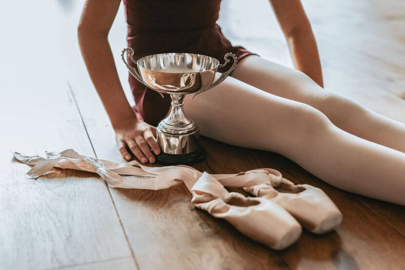 A teen ballet dancer sits gracefully on the floor, a bright trophy by her side and pointe shoes resting nearby, capturing a moment of youthful achievement and dedication.