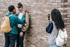 Three teens by a brick wall; one is being pressured and recorded, emphasizing the need for confidence-building support. A therapist in Scotch Plains NJ can help. 