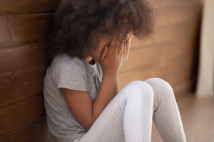 A young girl sits against a wooden wall with her knees drawn up, covering her face with her hands. She appears to be upset or overwhelmed, representing mental health concerns that sandplay therapy in Branchburg, NJ can address. Learn more about how a trauma therapist in Scotch Plains, NJ can offer support by searching for sand therapy in NJ and other services to support your child.