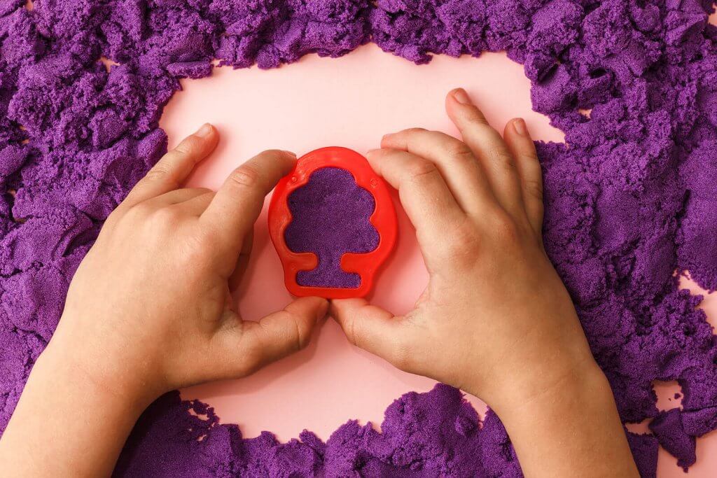 A close-up of a child's hands pressing a red plastic mold into purple kinetic sand. The child is shaping the sand, engaging in sensory play. Learn more about the power of play and how sandplay therapy in Branchburg, NJ can offer support. Search for child therapy in Somerville and contact a trauma therapist in Scotch Plains, NJ for more info. 
