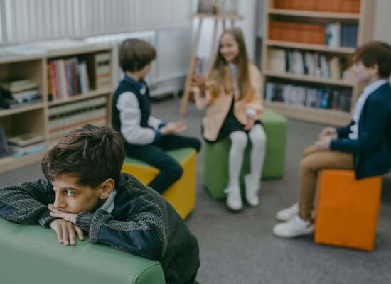 A young boy with dark hair and a sad expression leans on a green cushioned seat in a library or classroom setting. In the background, three other children are engaged in conversation. Learn more about the support child therapist in scotch plains, NJ can offer by searching for a selective mutism therapist. They can offer child therapy in New Jersey and other services.