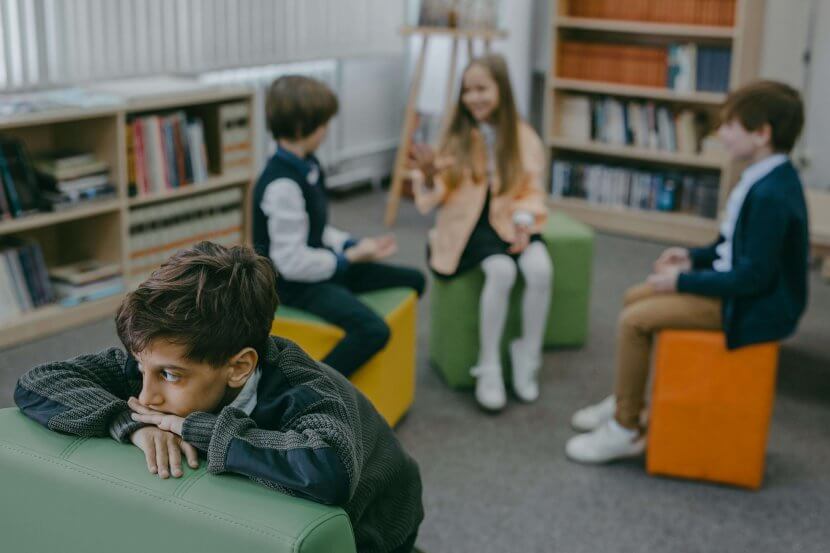 A young boy with dark hair and a sad expression leans on a green cushioned seat in a library or classroom setting. In the background, three other children are engaged in conversation. Learn more about the support child therapist in scotch plains, NJ can offer by searching for a selective mutism therapist. They can offer child therapy in New Jersey and other services.