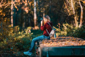 A young woman in a red hoodie and jeans sits on a weathered wooden structure covered in autumn leaves, holding a camera to her face. This could represent pursuing other hobbies after equestrian therapy in New Jersey. Learn more about how an EMDR therapist in Sommerville, NJ can help equestrians redefine themselves and overcome past trauma through EMDR therapy in Scotch Plains, NJ today. 